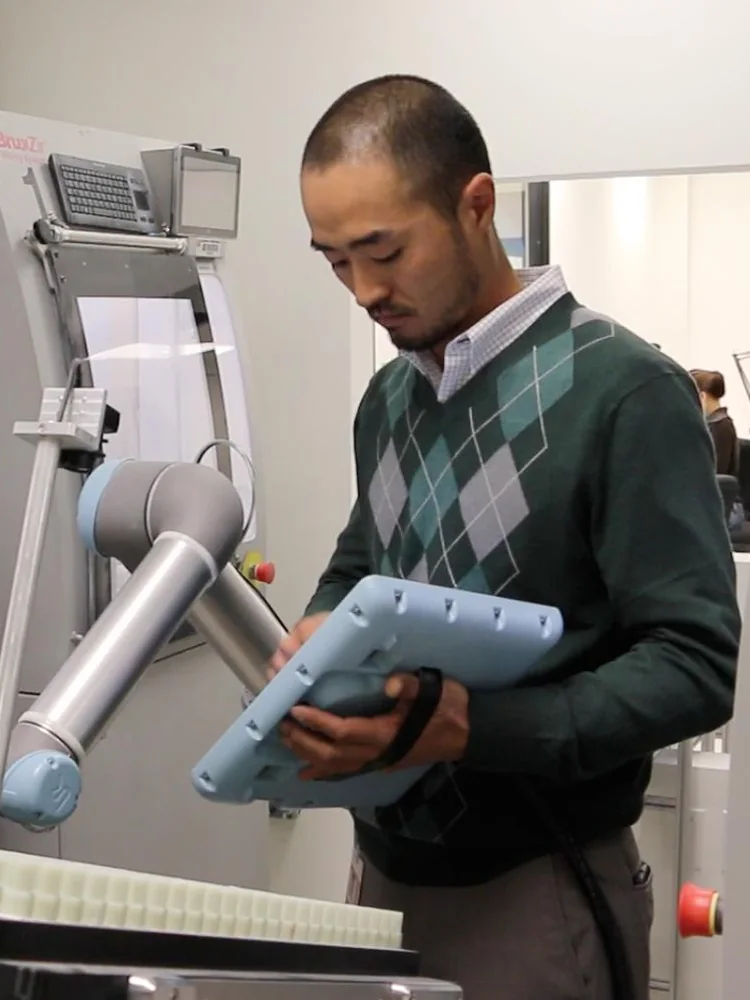 Técnico com o painel de programação ao lado de um robô colaborativo da Universal Robots, em frente às máquinas